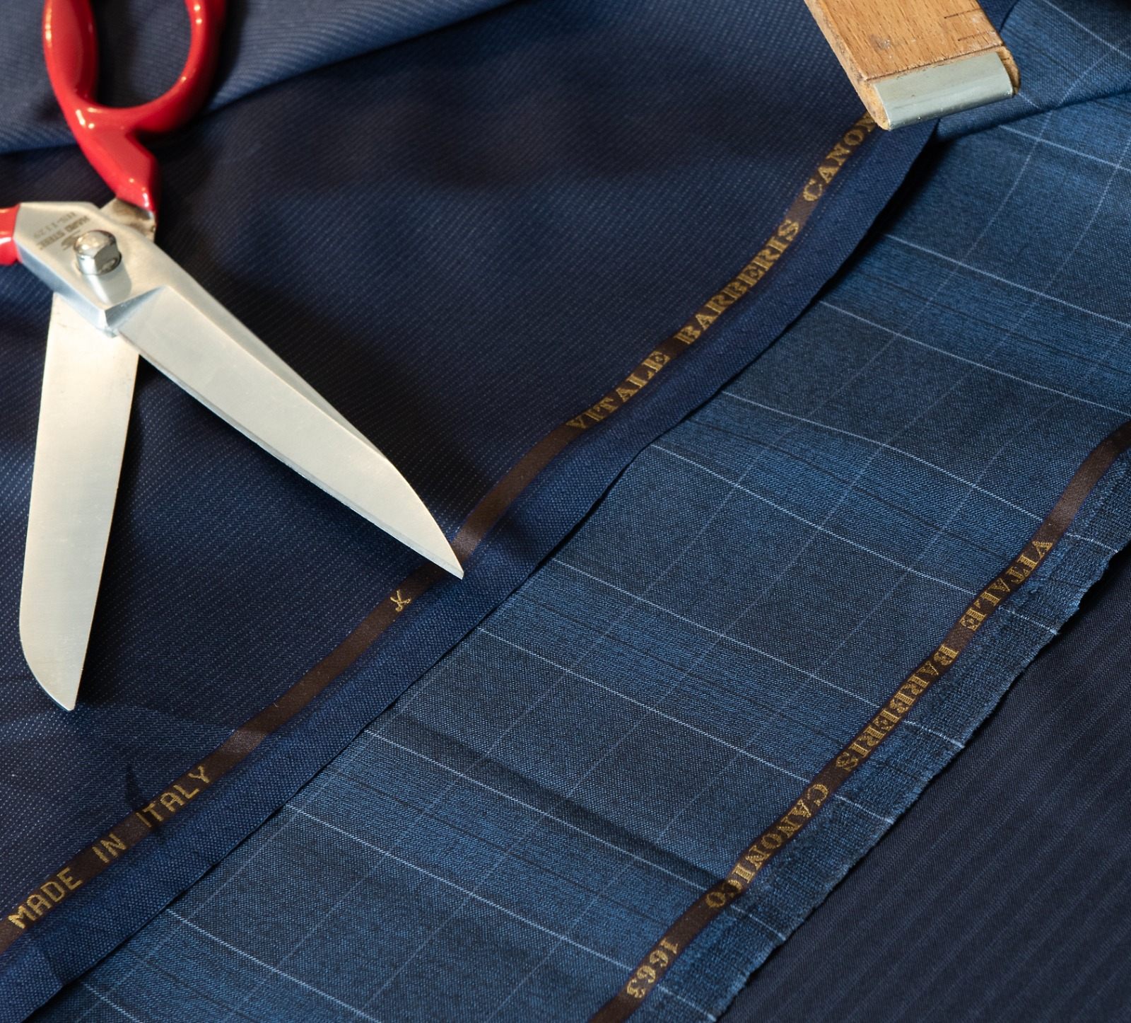 Üst üste serilmiş lacivert takım elbise kumaşları; kenar şeritlerinde altın renkli “VITALE BARBERIS CANONICO 1663” ve “MADE IN ITALY” yazıları okunuyor, üzerlerinde kırmızı saplı terzi makası ve ahşap metre
